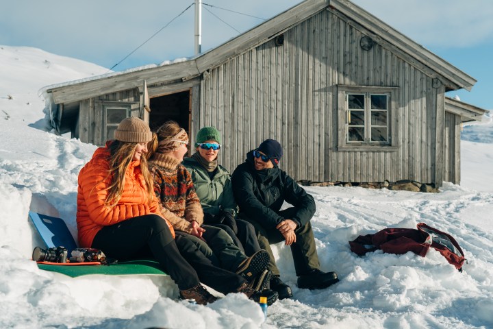 Four people sitting in snow outside a wooden cabin, dressed in winter clothing, under a clear blue sky.