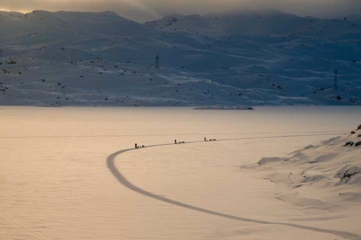 Sled dog team on snowy landscape with mountains in the background during sunset.