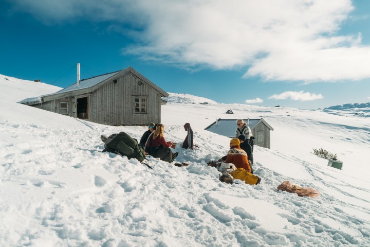 Group of people sitting on snow near huts, with a sunny blue sky.