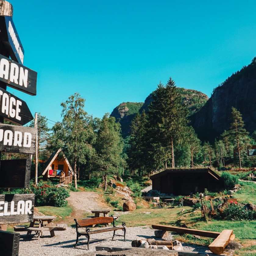 Rustic campsite with signs, benches, cabin, trees, and mountains under a clear sky.