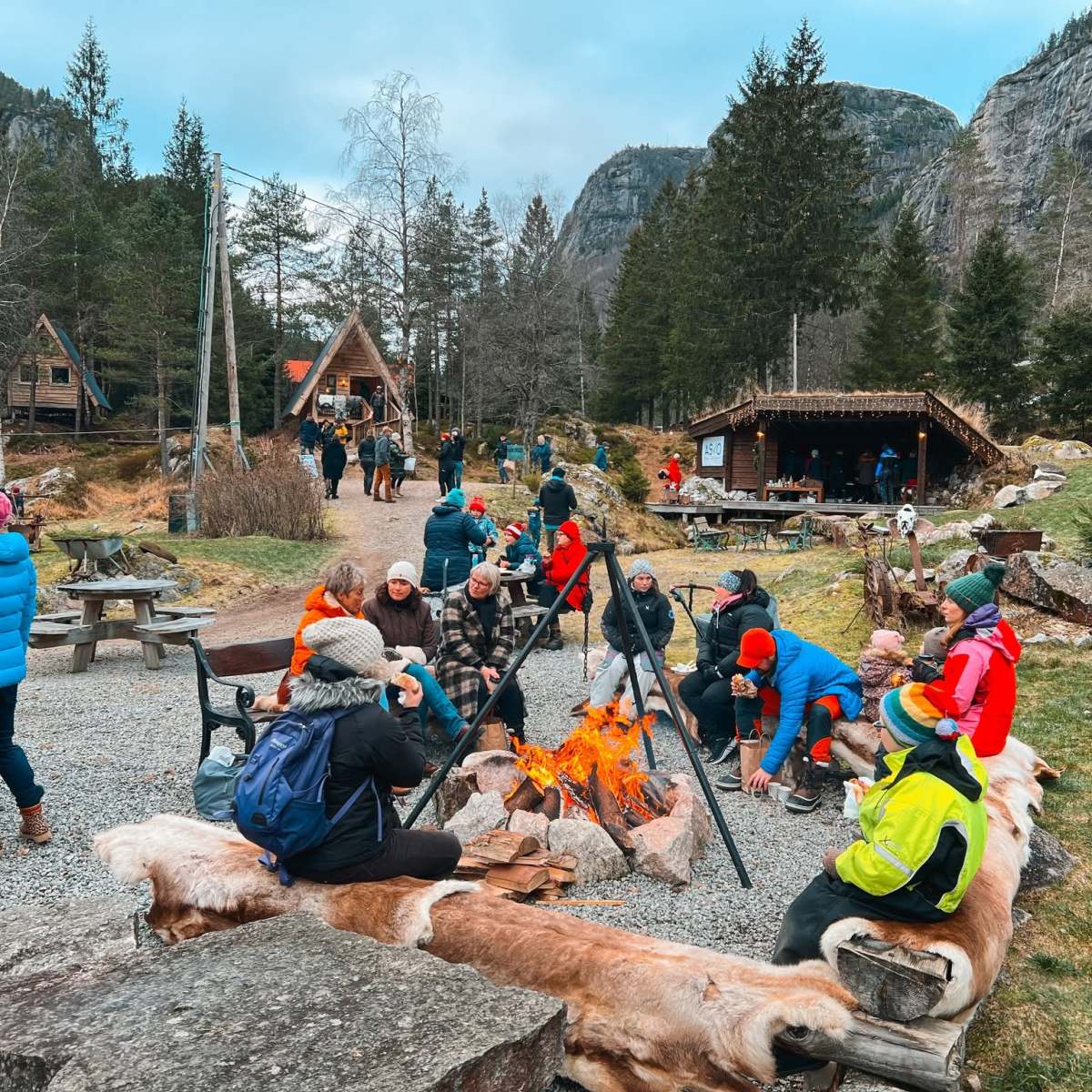 People in winter wear gathered around a campfire in a wooded outdoor area with cabins.