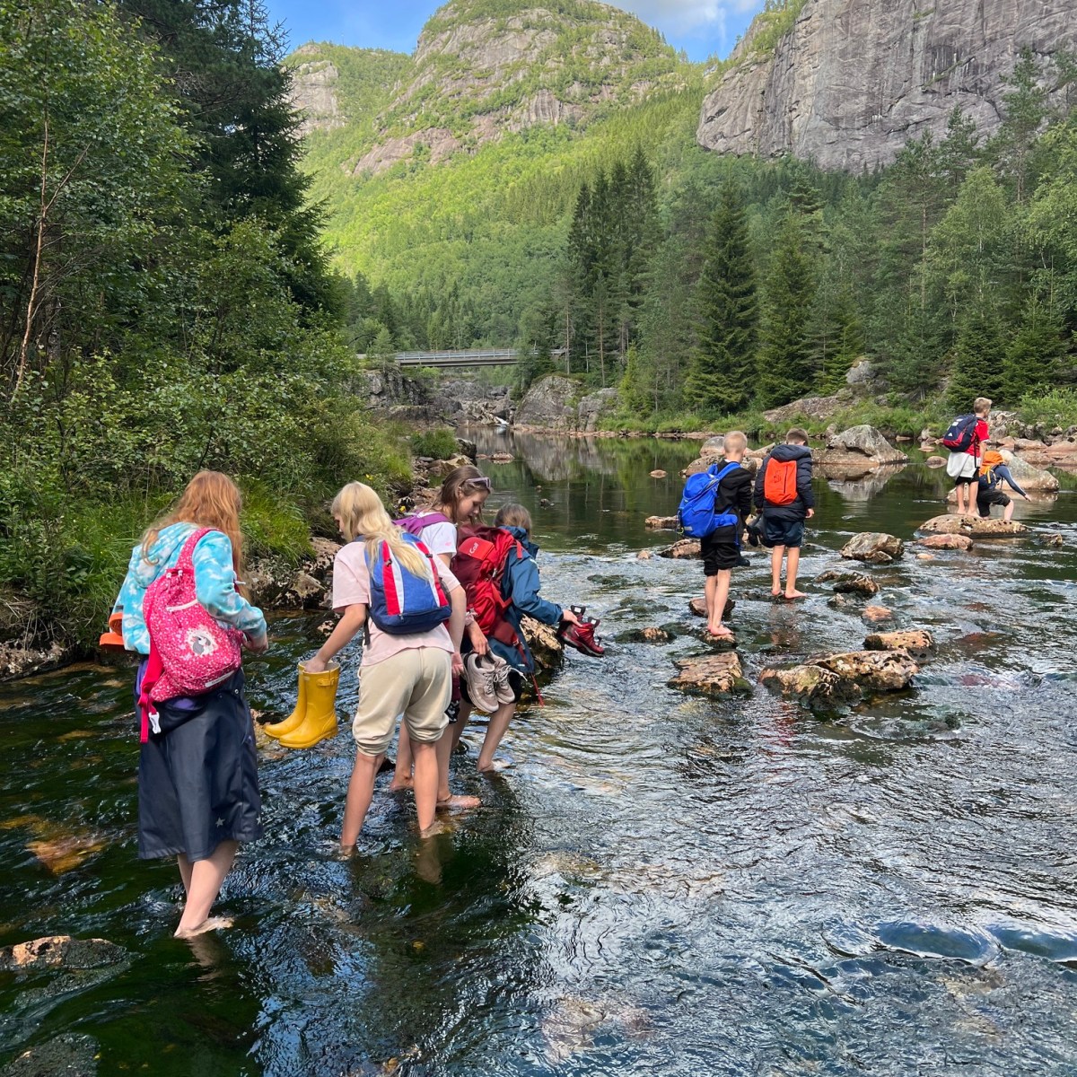Group of people crossing a shallow river surrounded by forest and mountains.