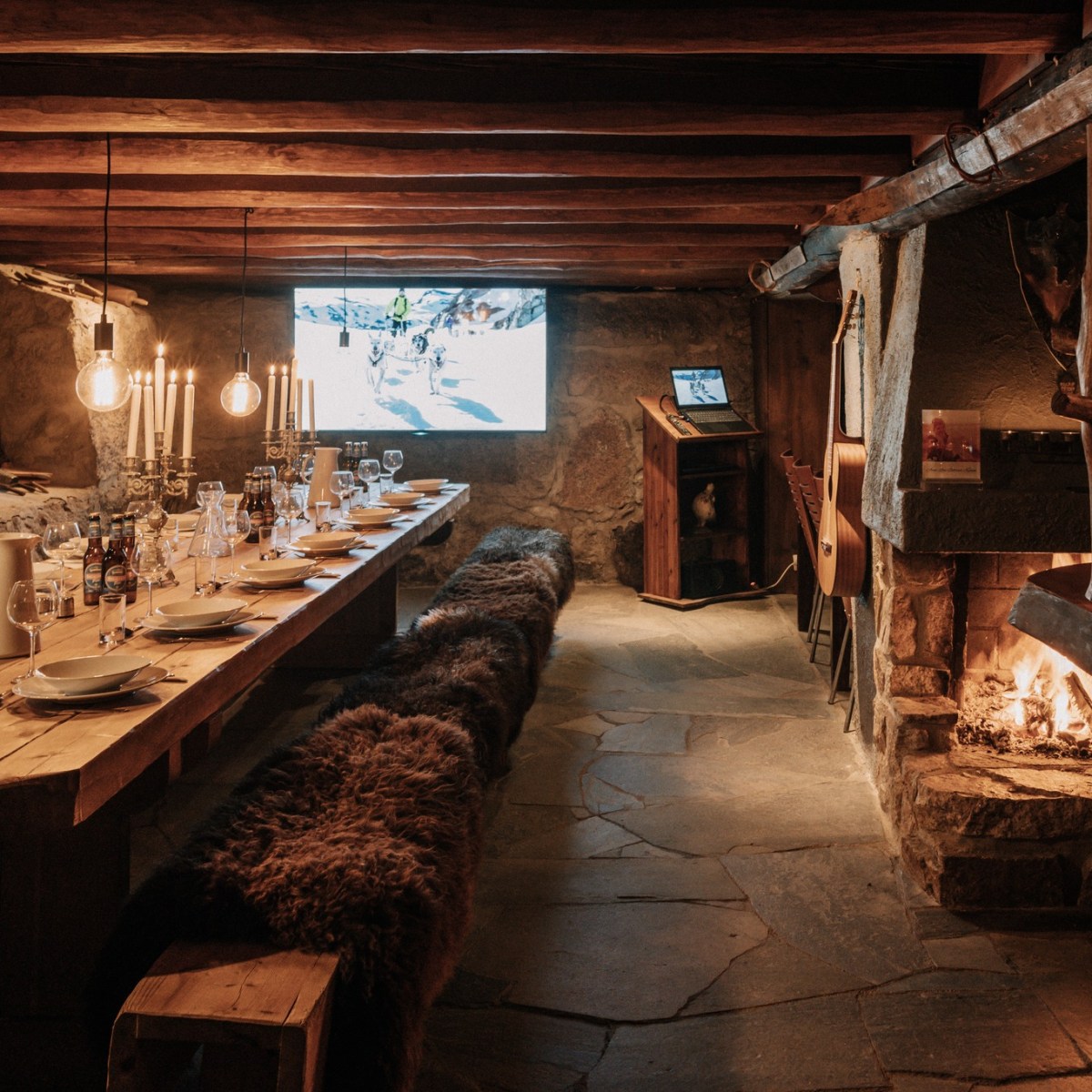 Cozy stone dining room with fireplace, long table, candles, and laptop displaying an image.