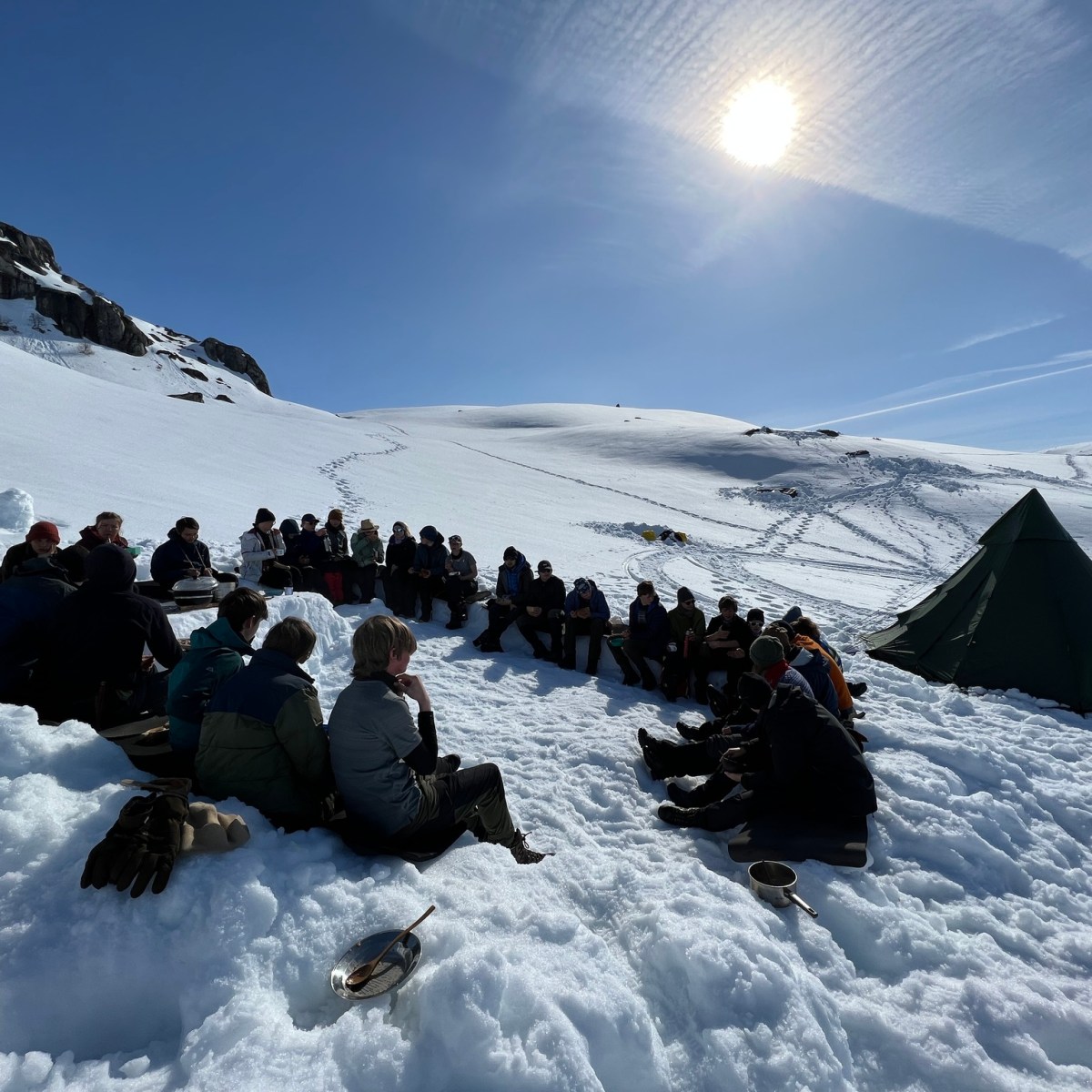 Group sitting in snow near a tent under a sunny sky with scattered clouds.