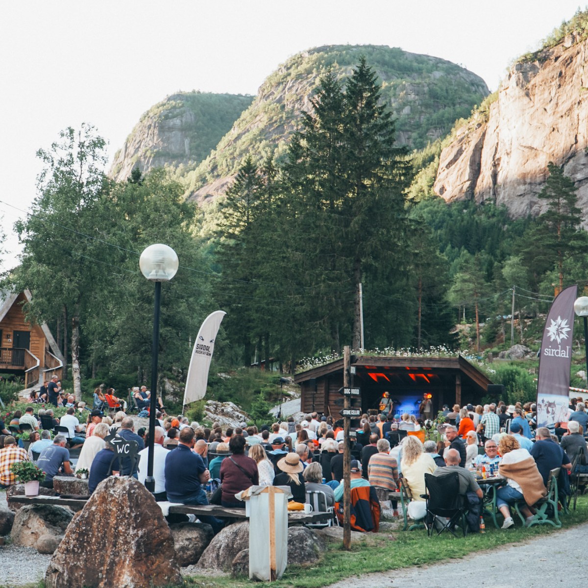 Outdoor concert with an audience in front of a stage set in a mountainous area.