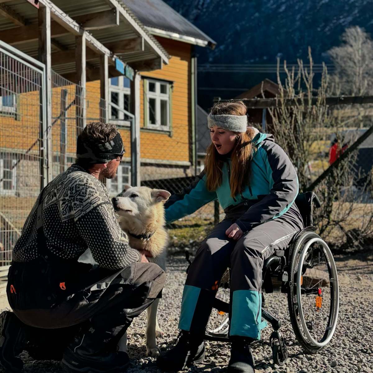 Woman in wheelchair and man pet a dog outdoors near a wooden building.