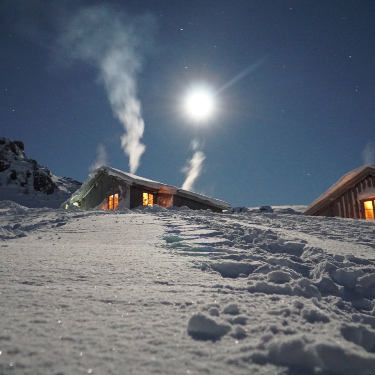 Snowy cabins with smoke under a bright moon in a wintery night scene.