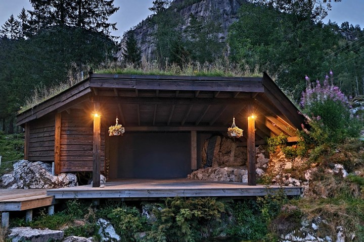 Wooden cabin with grass roof, lit by lanterns, near a pond and forested hill.